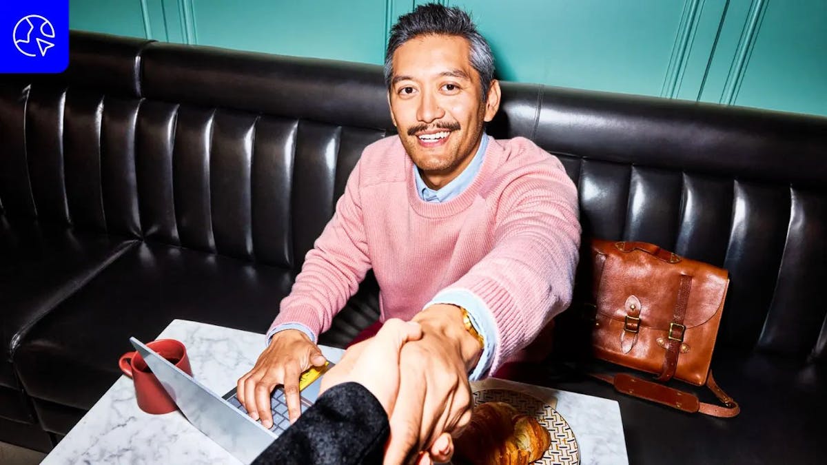 A man in a pink sweater smiles while shaking hands at a cafe table. He sits on a black bench next to a leather bag; a laptop, red cup, and pastry are on the table. The background features a textured teal wall