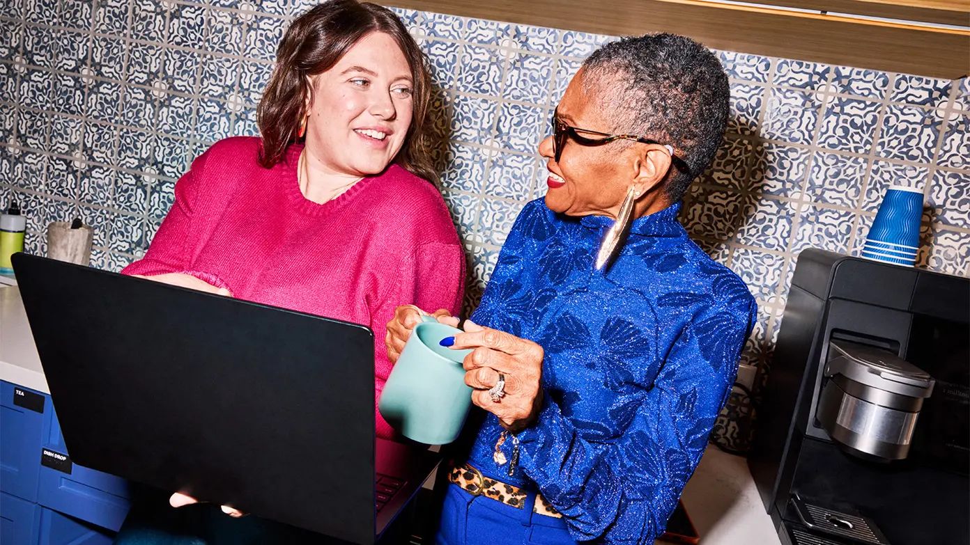 two woman looking at laptop standing in front of the office coffee machine