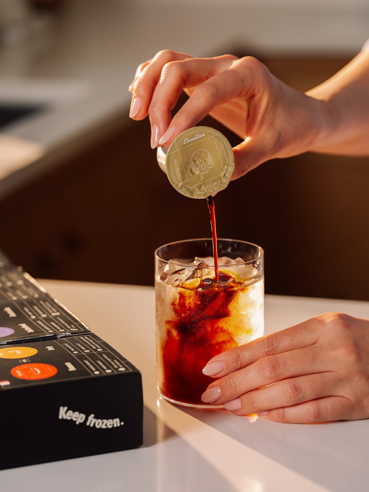 A Cometeer capsule pouring dark coffee over ice in a clear glass — no sugar, no milk