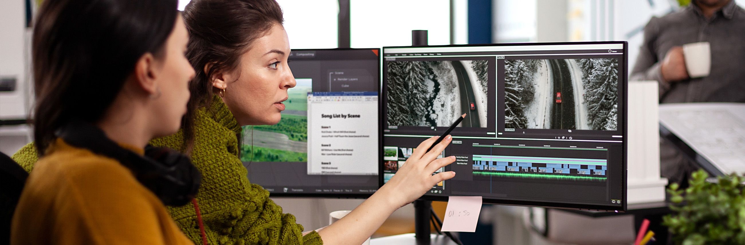 Two people working at a computer, with one pointing to a screen showing video footage and editing software.