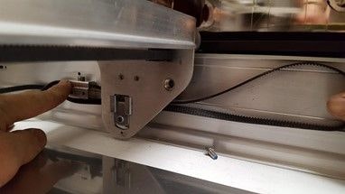 A finger holding the belt clamp to the side of the Glowforge and sliding it behind the side of the laser arm