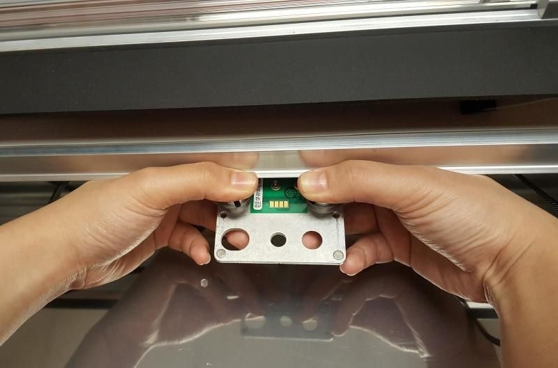 An image showing two hands grasping the front of the carriage plate that the Glowforge print head rests on.