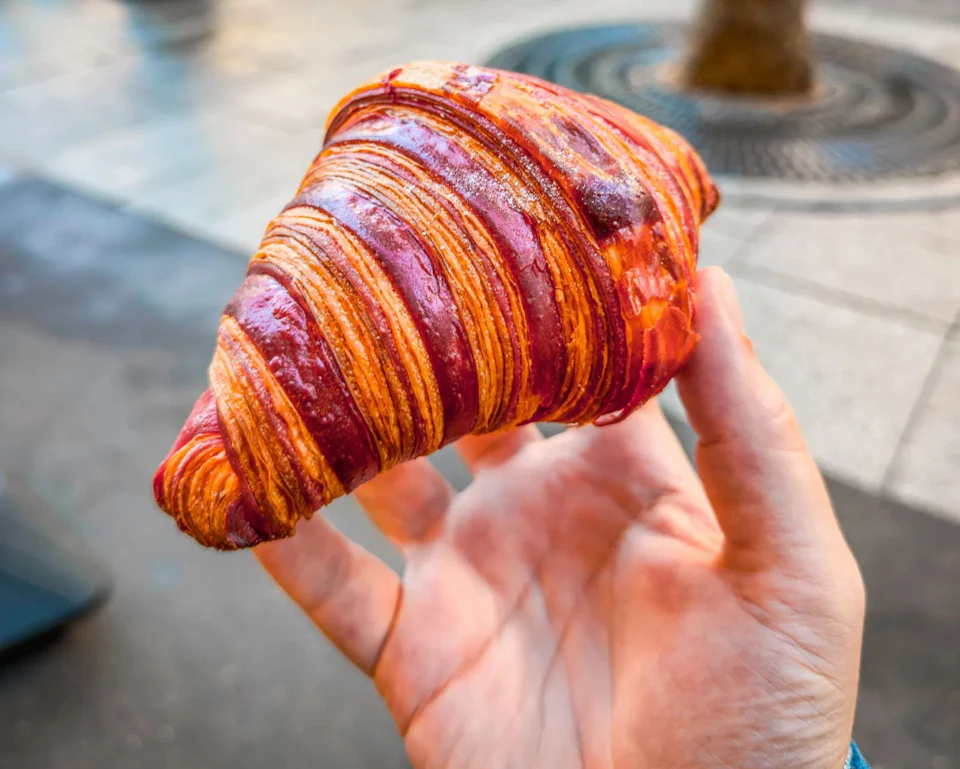 A hand holding a deep-red laminated croissant on a pavement