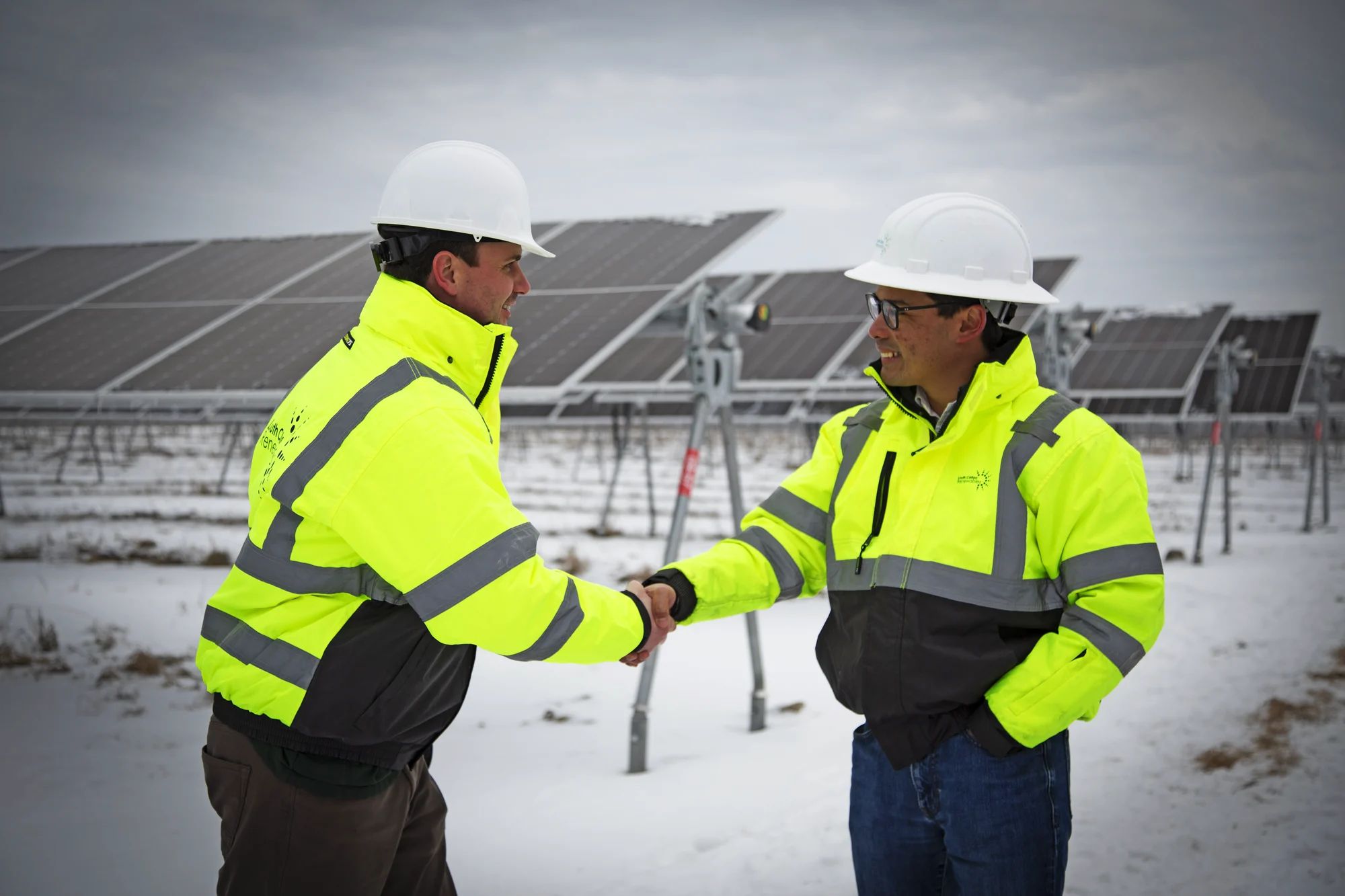 This image shows two individuals shaking hands in an outdoor setting covered with snow. Both individuals are wearing high-visibility jackets with reflective stripes and white hard hats. The background features a solar panel array, indicating that the location is likely a solar farm. The individuals appear to be engaged in a professional interaction, possibly related to the solar energy industry. The sky is overcast, and the ground is uniformly covered with snow.