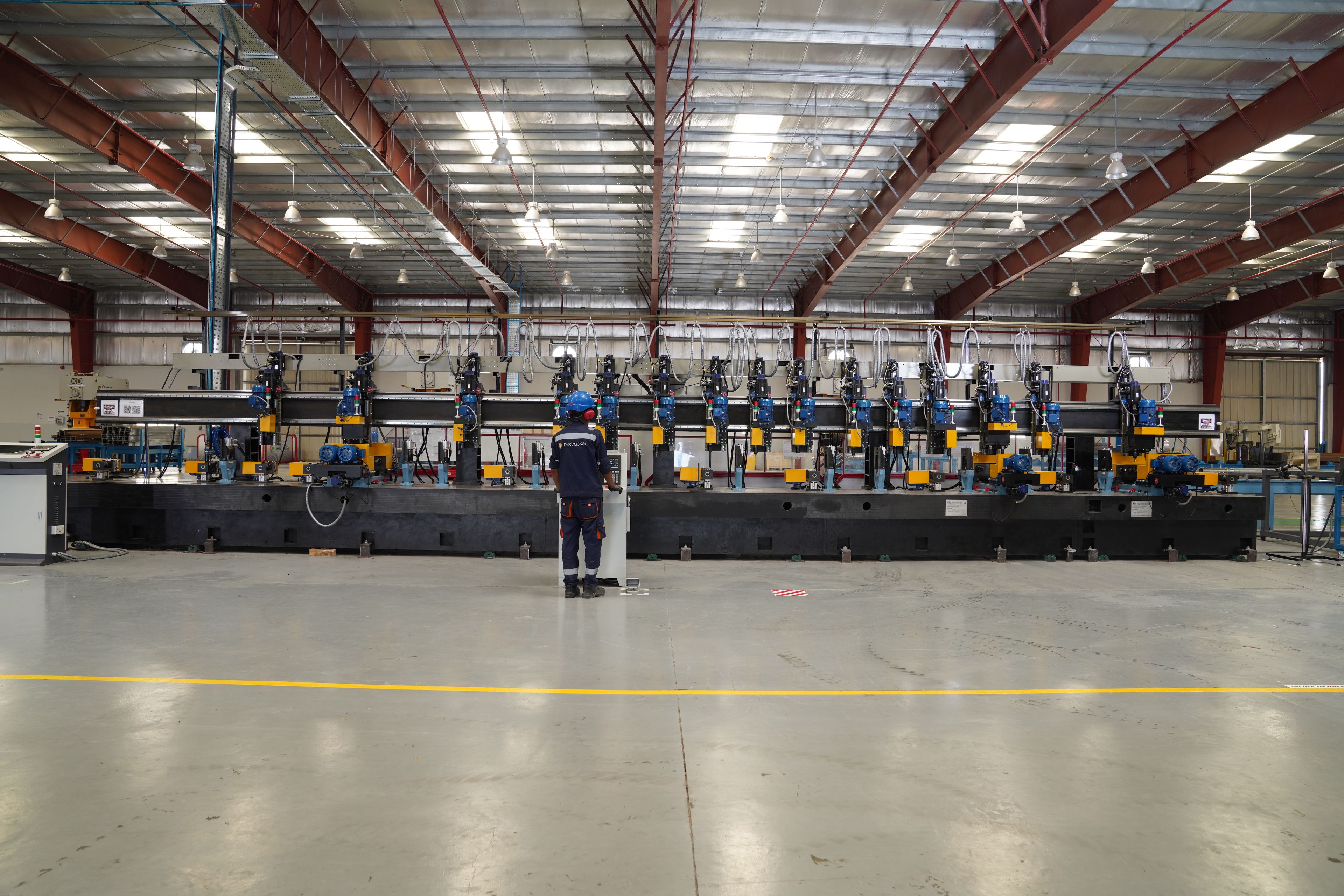 This image shows a large industrial facility with a high ceiling supported by metal beams. The floor is marked with a yellow line running horizontally across the bottom. A person wearing a blue helmet and a high-visibility vest is standing in front of a long, complex machine that spans the width of the facility. The machine consists of multiple units with blue, yellow, and black components, and it appears to be used for manufacturing or processing materials. Overhead, there are rows of lights suspended from the ceiling, providing illumination for the workspace.