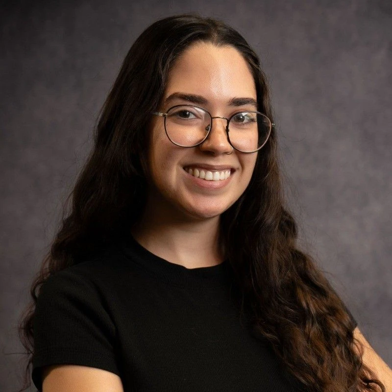 Professional headshot of a smiling young woman with long dark hair and glasses against a grey studio background.​​ 