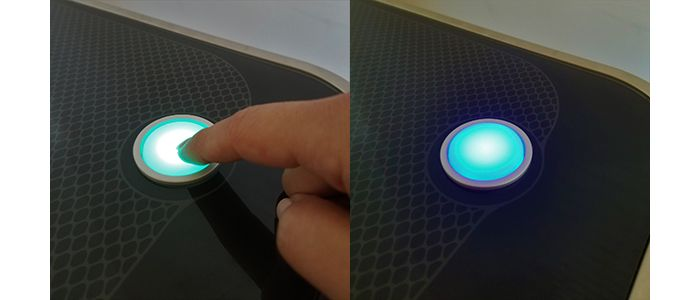 A finger holding down the Glowforge button when teal, and then the button turning blue after being released