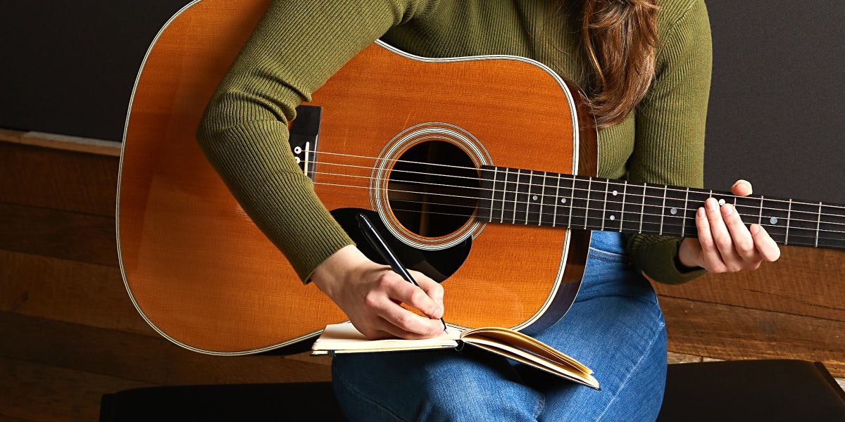 Person holding an acoustic guitar, and writing in a notebook