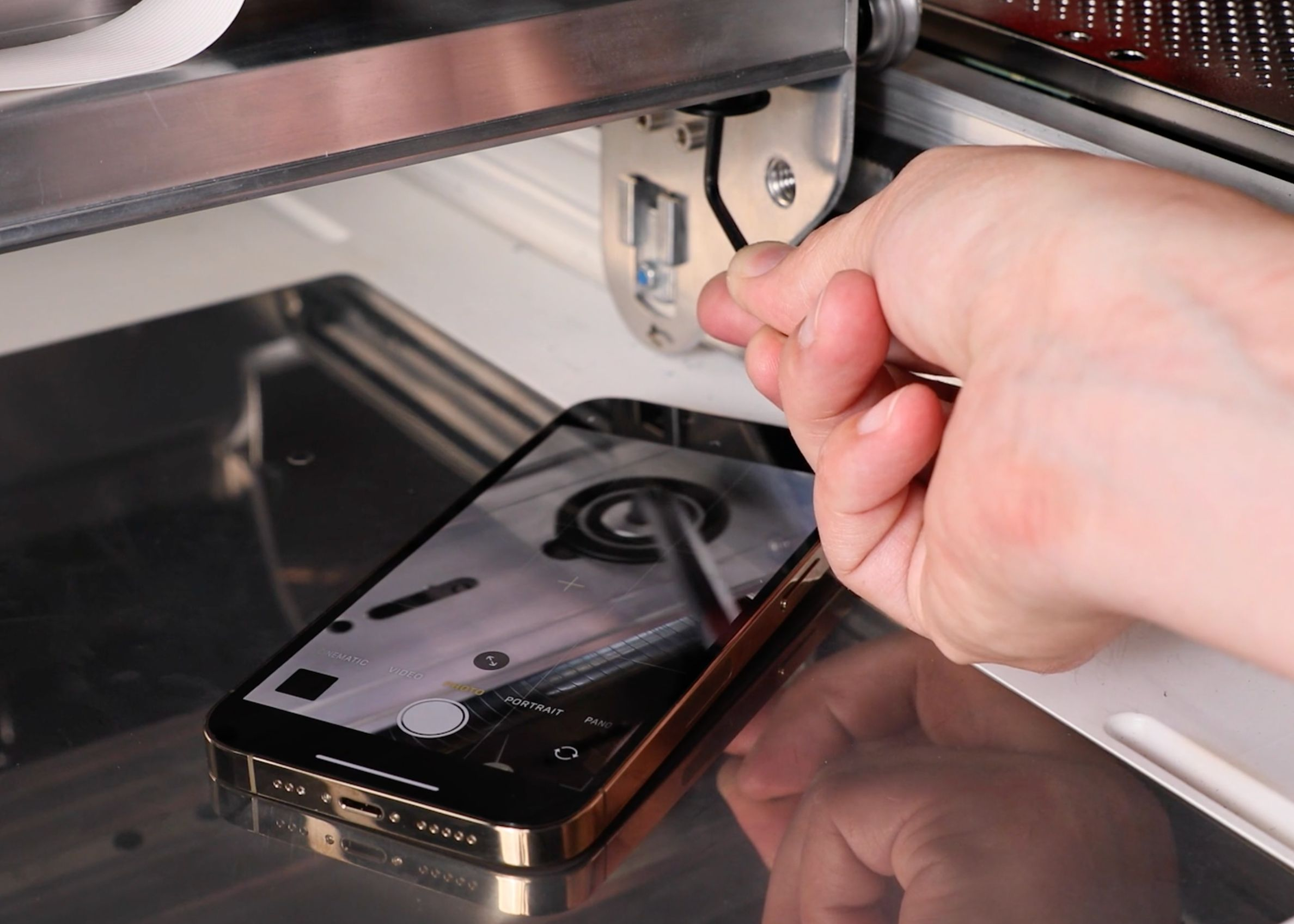 A hand holding a hex key inserted into the pulley screw. A phone in selfie mode is in the bed of the Glowforge, pointed at the pulley
