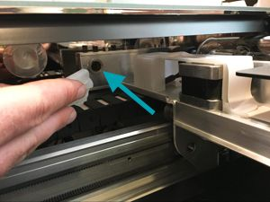 A hand holding a lens wipe and cleaning the window located inside the left-hand portion of the Glowforge