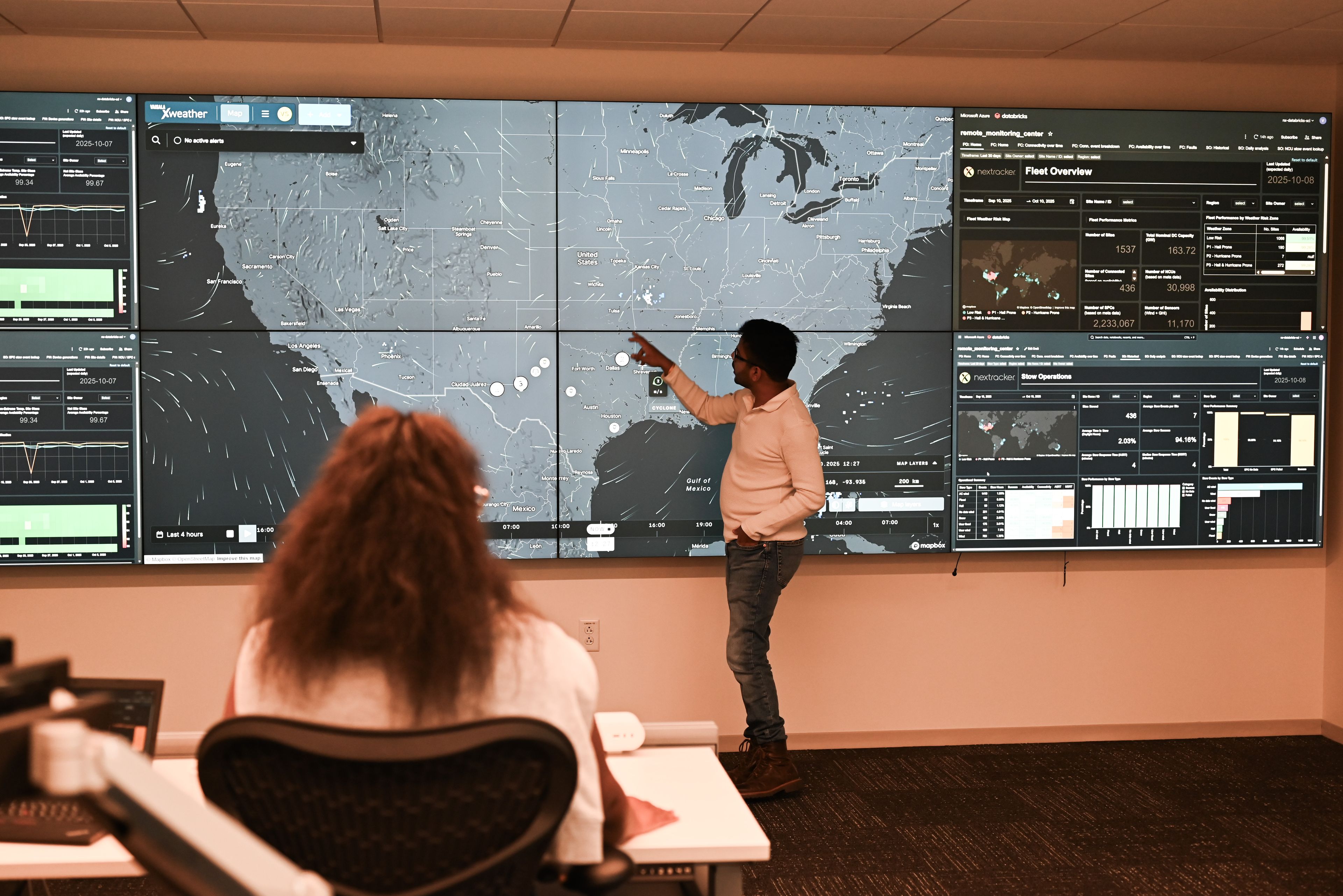 This image shows a control room with a large wall-mounted display showing various data visualizations and maps. Two individuals are present, one standing and pointing at the display while the other is seated in front of a desk with computer monitors. The display includes multiple sections with different types of data, such as weather maps, fleet overviews, and operational statistics. The standing individual appears to be explaining or discussing the information on the display.
