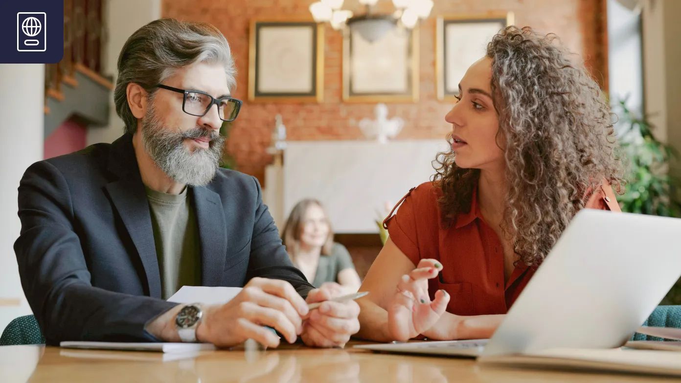 Un uomo con la barba grigia e una donna con i capelli ricci si siedono a un tavolo in un ufficio luminoso, impegnato in una discussione seria su documenti e un laptop.