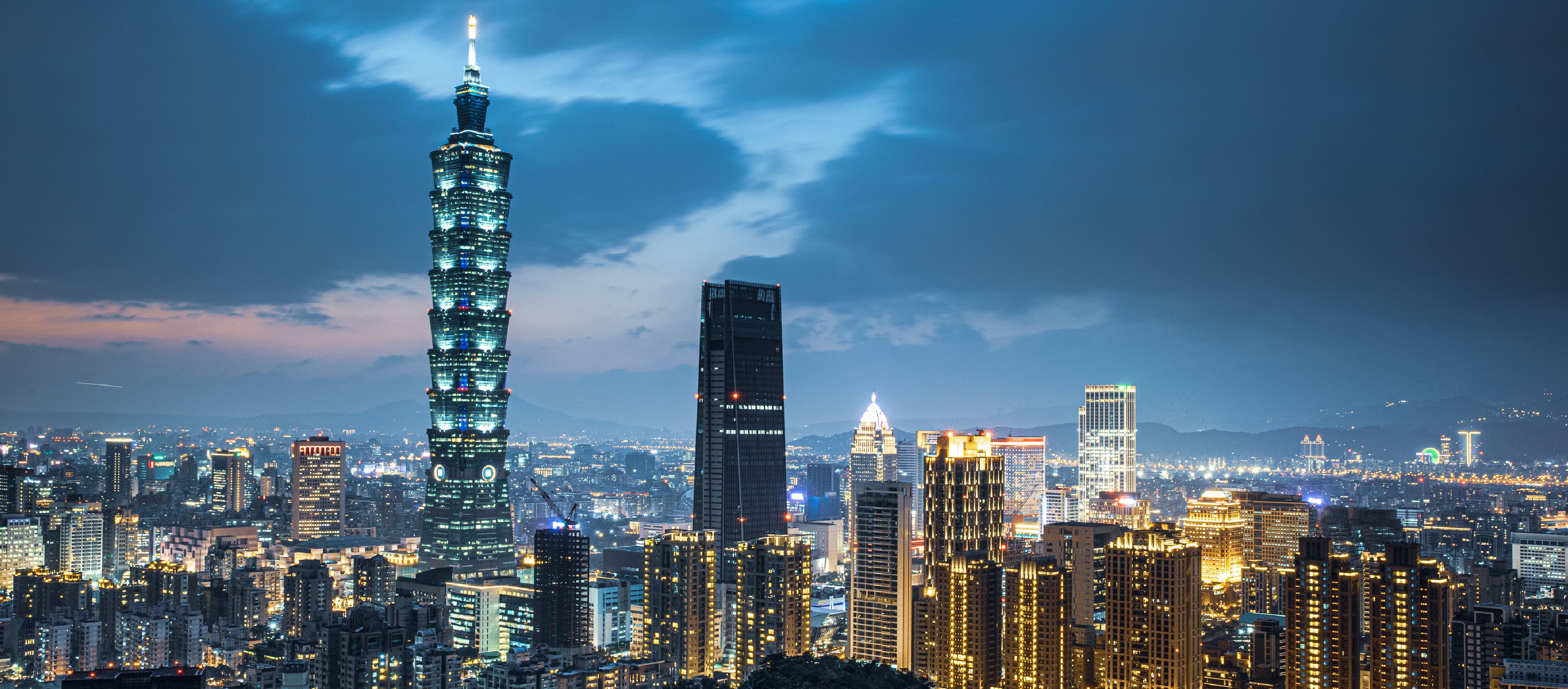 This image shows a vibrant cityscape at dusk or dawn, dominated by a prominent, tall, multi-tiered building with a spire at the top center.