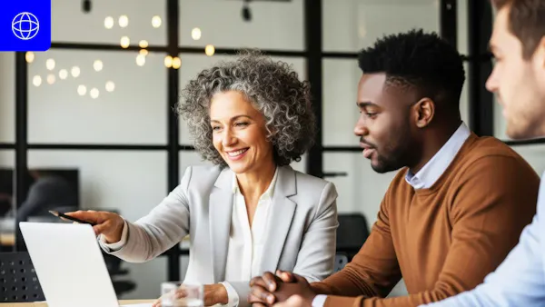 A diverse group of three professionals in a modern office setting looking at a laptop. A woman with gray curly hair is pointing at the screen while two male colleagues watch and listen. A blue globe icon is visible in the top left corner.