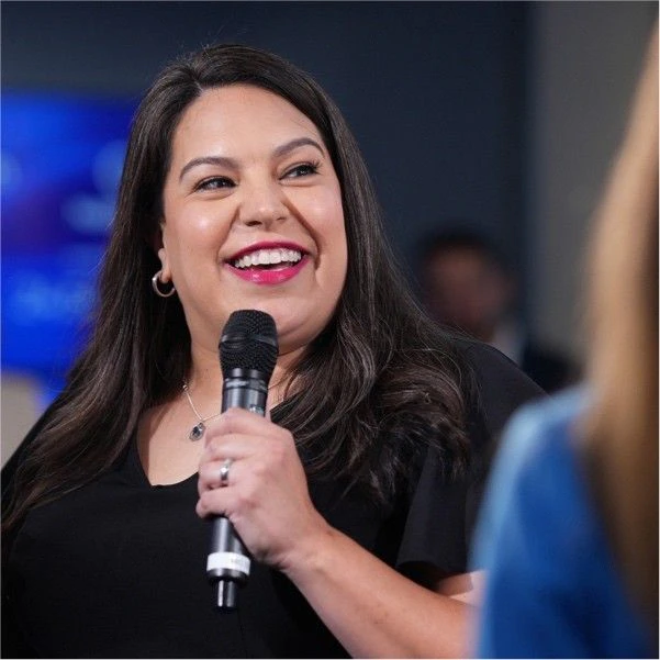 Smiling woman with dark hair holding a microphone and speaking at a panel discussion​​ 